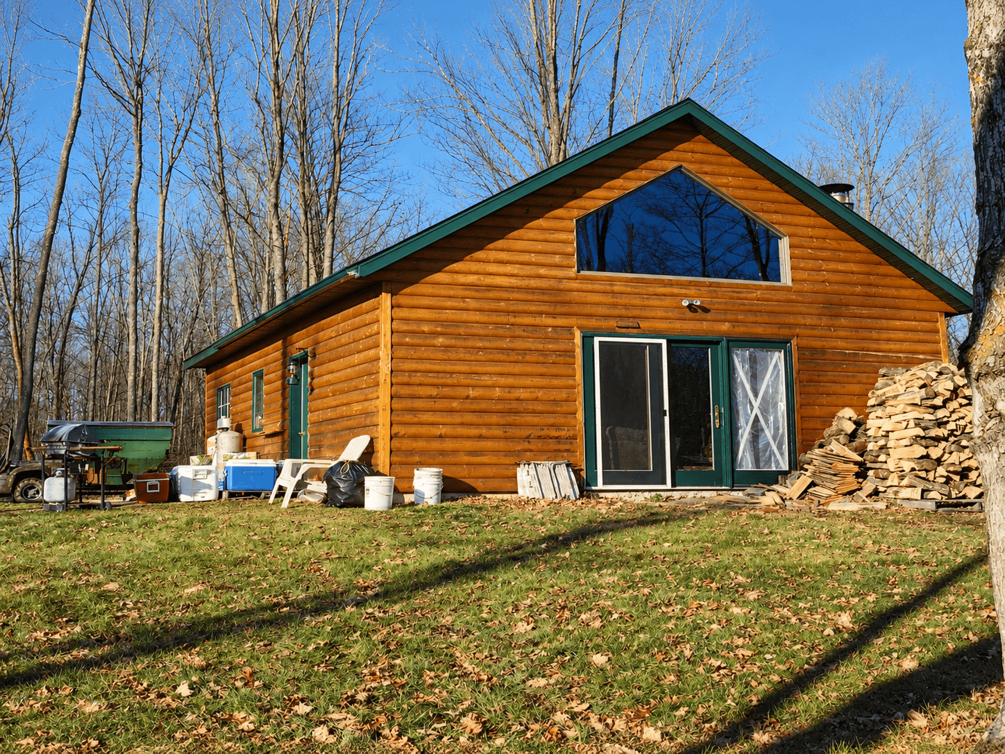 Log cabin with green trim, sliding glass door, stacked firewood and coolers on lawn