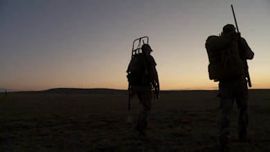 Two silhouetted hunters walking across open New Mexico plains at sunrise; one carries a rifle