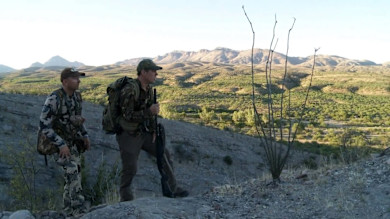 Two hunters on rocky ridge overlooking desert valley; one leans on a shotgun.