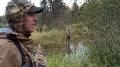 Hunter in camo hooded jacket beside creek, speaking; another hunter wades with rifle