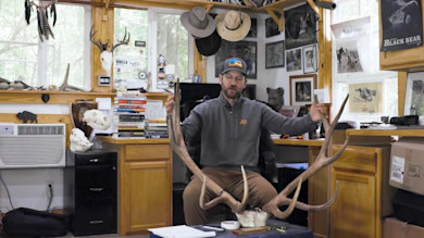 Man seated in wood-paneled office holding large elk antlers; poster "Black Bear", sticker "RINELLA"