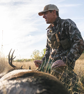 Hunter kneeling beside a buck with antlers in grassy field, wearing camo jacket and cap