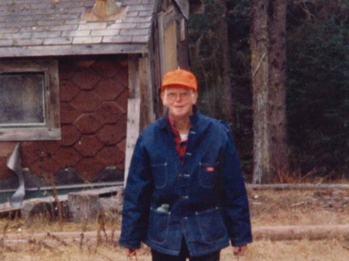 Older man in orange cap and glasses wearing a blue jacket standing before a small wooden cabin