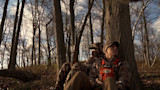 Two hunters sitting against a tree in bare woods; one holds a rifle upright, other wears orange harness