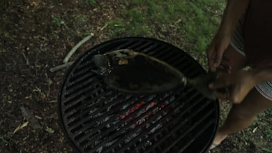 Whole fish held over round charcoal grill with glowing embers; person's hands and legs visible
