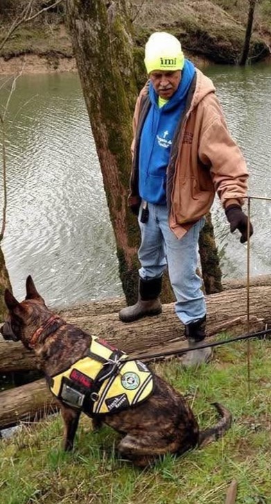 Man in neon beanie labeled 'imi' and blue hoodie standing on riverbank with working dog in yellow vest