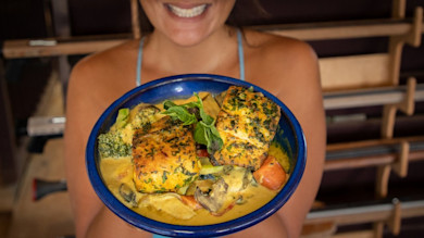 Blue bowl of fish curry with seared fish, vegetables, and basil held by smiling woman