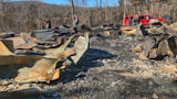 Charred cabin ruins of twisted corrugated metal and ash, three men by red SUV