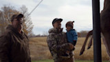 Man in camouflage jacket holding child beside hanging deer; another man laughs in field
