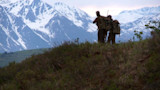 Two hunters with backpacks and rifles standing on a ridge overlooking snow-capped mountains