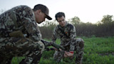Two hunters in camouflage kneel in a field, examining an arrow with red fletching