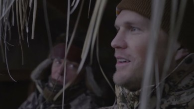 Two hunters in a grass-lined duck blind, right man in beanie speaking