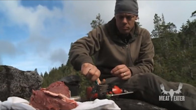Hunter cooking venison loin on portable stove atop rock, MEAT EATER logo