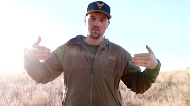 Hunter demonstrating pinch point, hands spaced, brown jacket with orange logo in grassy field