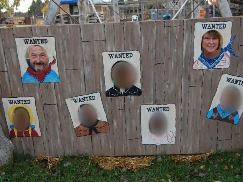 WANTED posters painted on wooden board with people posing through face cutouts; some faces blurred