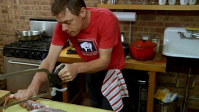 Man in red shirt sawing bone-in meat with butcher's saw at kitchen counter
