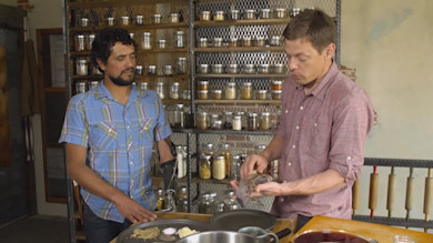 Two men, one with a prosthetic arm, preparing smoked meat in a pantry kitchen with shelves of spice jars