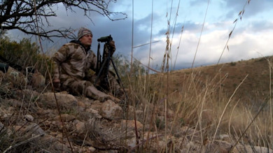 Hunter with spotting scope on tripod seated on rocky hillside in camouflage under cloudy sky