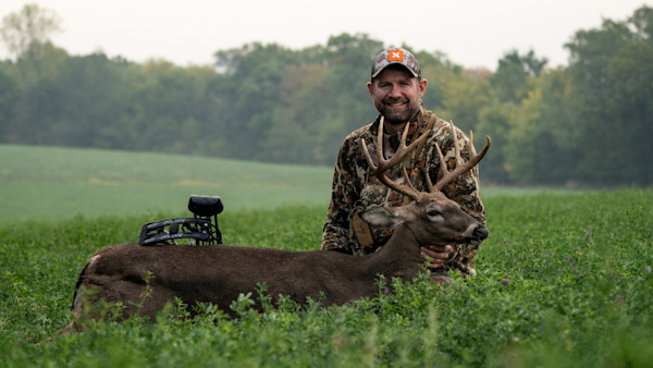 Hunter kneeling in green field holding a large ten-point buck with a compound bow