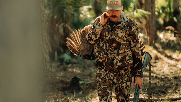 Hunter carrying turkey and shotgun in camouflage, cap reads YETI