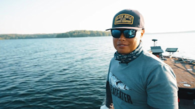 Angler on a boat at a lake wearing a cap reading "BLACK RIFLE COFFEE COMPANY" and a "MEATEATER" shirt