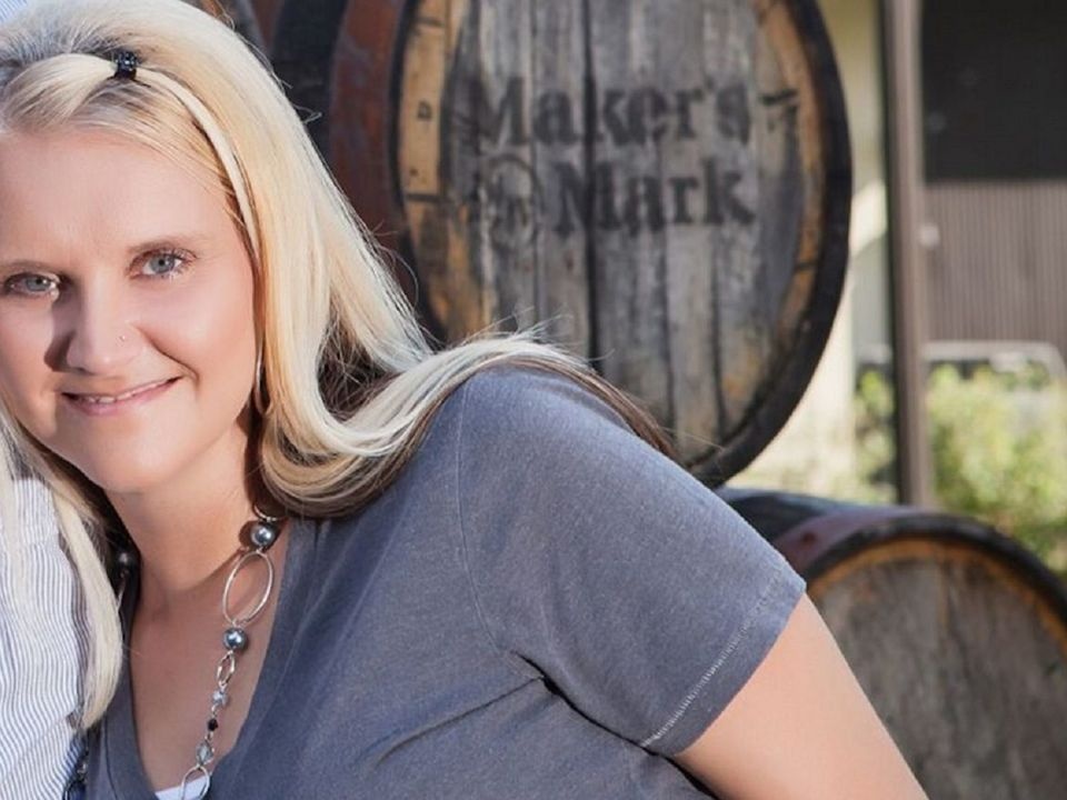 Blonde woman smiling in gray shirt and necklace; barrels behind read Maker's Mark