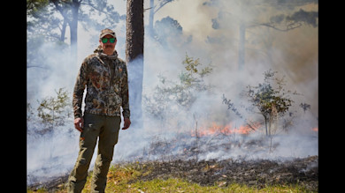 Hunter near controlled grass fire wearing camouflage and green mirrored sunglasses