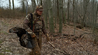 Hunter in camouflage walking through leafless woods carrying a harvested turkey and shotgun