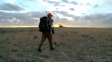 Hunter carrying rifle and large backpack walking across grassy prairie at sunrise
