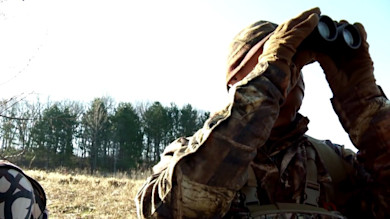 Hunter in camouflage and gloves looking through binoculars in a grassy field