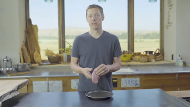 Steven Rinella holding raw pastrami slice over black plate in kitchen with window view