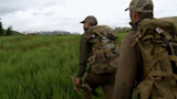 Two hunters with rifles and backpacks walking through tall grass toward snow-capped mountains