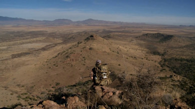Hunter with backpack sits on rocky ridge overlooking arid Arizona valley
