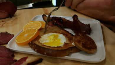 Toast with egg cooked in the center, orange slices, bacon strips and sausage on a white plate; hand holding fork.