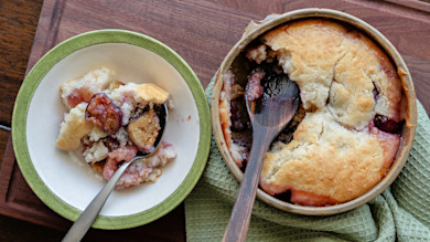 Fig cobbler with biscuit topping in baking dish, wooden spoon and a serving in bowl on board