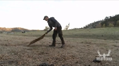 Man dragging tarp across dry field, MEAT EATER logo bottom right
