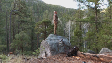 Hunter standing on boulder in pine forest with pack and rifle leaning against rock