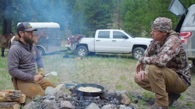 Two hunters kneel by a campfire cooking in a cast-iron skillet, truck and horse trailer behind