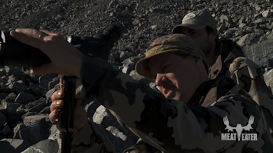 Two hunters adjusting a spotting scope on a rocky slope with "MEAT EATER" logo