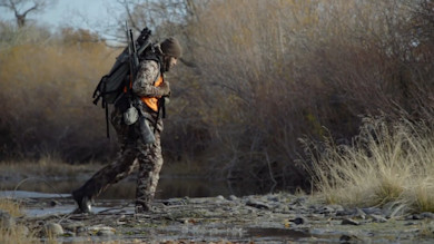 Hunter in camouflage and orange vest crosses shallow stream carrying backpack and rifle