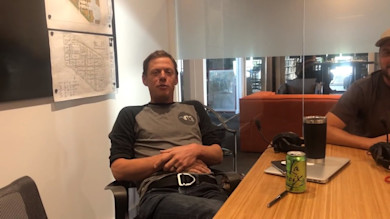 Man seated at table with podcast microphone, laptop, headphones and a LaCroix can