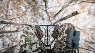 View from tree stand: camo-clad legs and binoculars over snowy forest floor