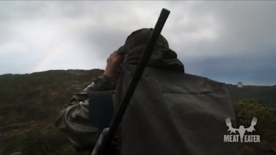 Hunter in rain jacket with slung rifle using binoculars on ridge with MEAT EATER logo