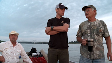 Three men on a boat by a river bridge; center wearing YETI cap and black First Lite shirt; right holding travel mug