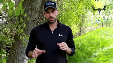 Man by tree in grassy field wearing black Under Armour top and cap reading "Wilderness Athlete"