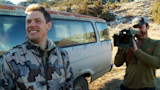 Smiling hunter in camouflage jacket beside van; cameraman filming with shoulder camera
