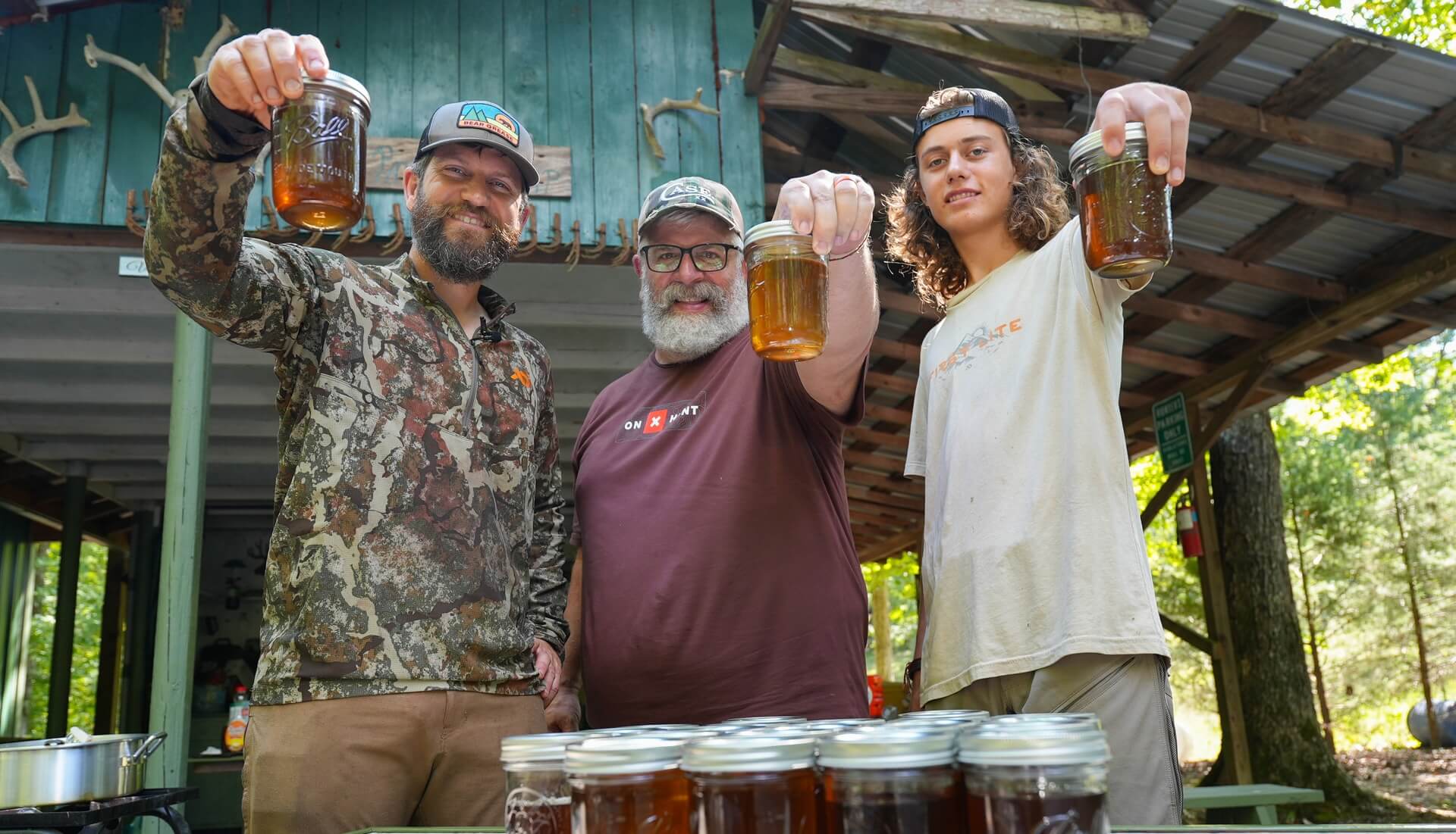 Three men holding mason jars of amber liquid; left man wearing Bear Grease hat