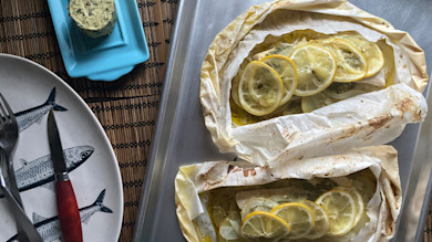 Two lemon-topped fish fillets baked in parchment on a sheet pan, herb butter and fish-print plate beside