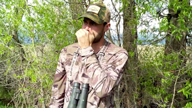 Hunter in camouflage cupping hand to mouth to make elk mew, binoculars strap reads UNDER ARMOUR