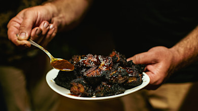 Smoky burnt ends on a white plate held by two hands as a spoon drizzles barbecue sauce